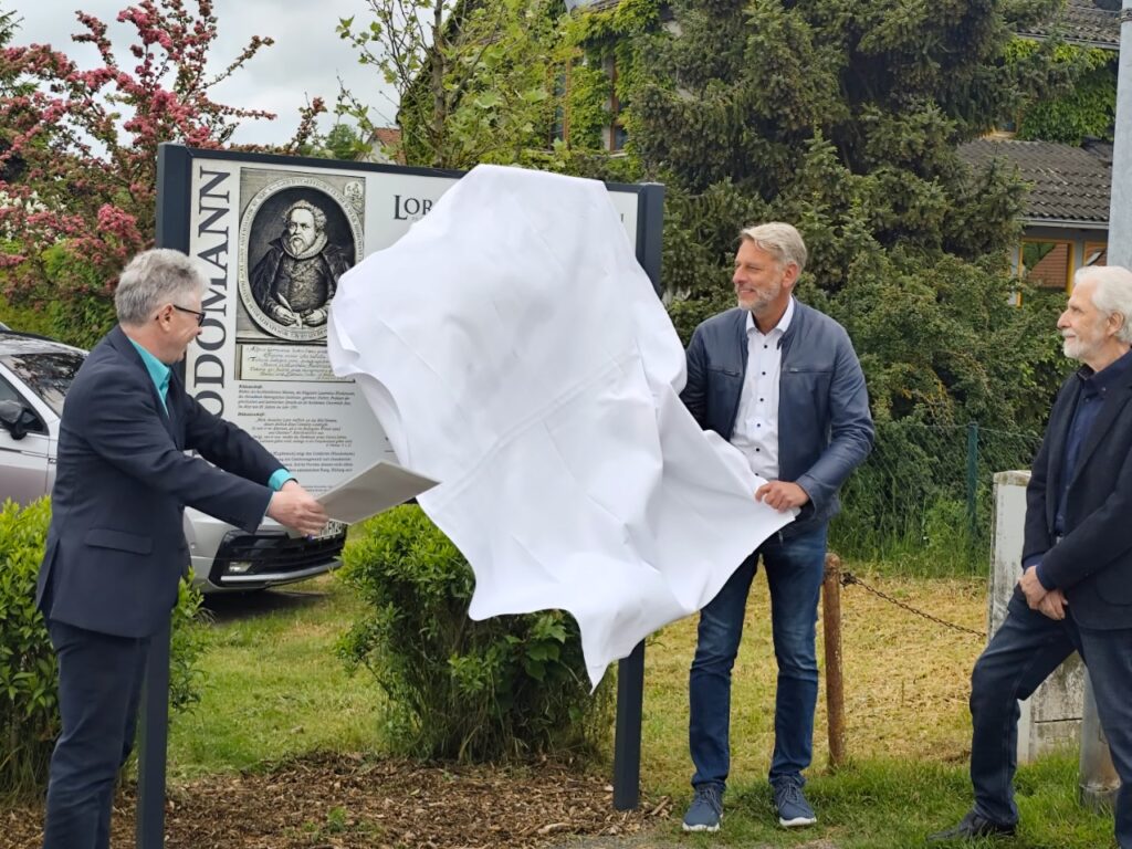 Drei Männer enthüllen vor einer großen Schautafel ein weißes Tuch. Links ein Herr mit grauen Haaren und Brille (soweno-Heimleiter Dirk Erfurt), in der Mitte Bürgermeister Stephan Klante mit dunkler Jacke, rechts ein Herr mit weißem Bart. Im Hintergrund die neue Gedenktafel mit Porträt von Lorenz Rhodomann. Standort: Rhodomannstraße in Niedersachswerfen.