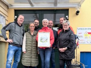 Sieben Personen stehen vor dem Eingang des Kindergartens Regenbogen in Neustadt. Die Frau in der Mitte tr&auml;gt eine rote Jacke und h&auml;lt einen gerahmten Gutschein in den H&auml;nden. Der Gutschein zeigt ein Bild und die Aufschrift: Sonnensegel vom Ortschaftsrat Neustadt, Holzgestell gesponsert von Massivholz-M&ouml;bel Thomas K&uuml;hn. Im Hintergrund ist die gr&uuml;ne Eingangst&uuml;r und ein Plakat mit der Aufschrift &bdquo;Leitbild der Kita" zu sehen. Die Gruppe l&auml;chelt in die Kamera.