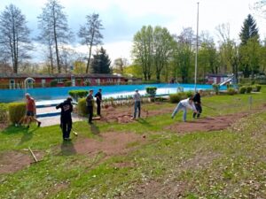 Mehrere Helferinnen und Helfer arbeiten mit Schaufeln und Besen an einem Erdstreifen direkt am leeren Schwimmbecken des Waldbades. Im Hintergrund ist das blaue, leere Becken zu sehen, dazu die roten Umkleideh&auml;uschen und B&auml;ume mit frischem Fr&uuml;hlingsgr&uuml;n. Mehrere Jugendliche aus dem Jugendclub Neustadt packen mit an. Die Stimmung wirkt arbeitsam und zugewandt.