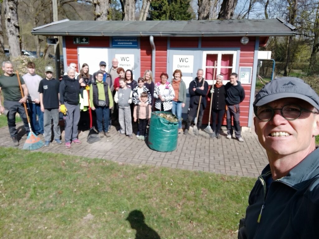 Eine größere Gruppe von Helferinnen und Helfern, darunter auch Kinder, steht in einer Reihe vor den roten Toilettenhäuschen des Waldbades. Viele halten Rechen und Besen. In der Mitte steht ein großer grüner Sack voller Laub. Im Vordergrund ist als Selfie der Fotograf zu sehen, der lächelnd in die Kamera blickt. Sonniger Frühlingstag mit klarem Licht.
