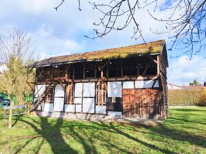 Ein kleines Fachwerkgeb&auml;ude mit verwittertem, bemoostem Dach steht auf einem Grundst&uuml;ck in Neustadt (Harz). Die wei&szlig;en Gefache zwischen dunklen Holzbalken sind noch gut erkennbar. Das Dach zeigt deutliche Sch&auml;den. Im Hintergrund sind Wohnh&auml;user und kahle B&auml;ume zu sehen. Das Foto wurde im Fr&uuml;hjahr aufgenommen.