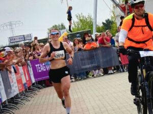 Eine L&auml;uferin mit der Startnummer 42010 erreicht den Zielbereich des 12. Harztorlaufs in Niedersachswerfen. Sie tr&auml;gt ein schwarzes Sport-Top, schwarze Shorts und eine verspiegelte Sportbrille. Sie l&auml;chelt. Hinter den Absperrgittern applaudieren Zuschauerinnen und Zuschauer. Rechts begleitet sie ein Helfer in oranger Warnweste auf einem Mountainbike. Im Hintergrund Werbebanner und ein TEAG-Schriftzug.