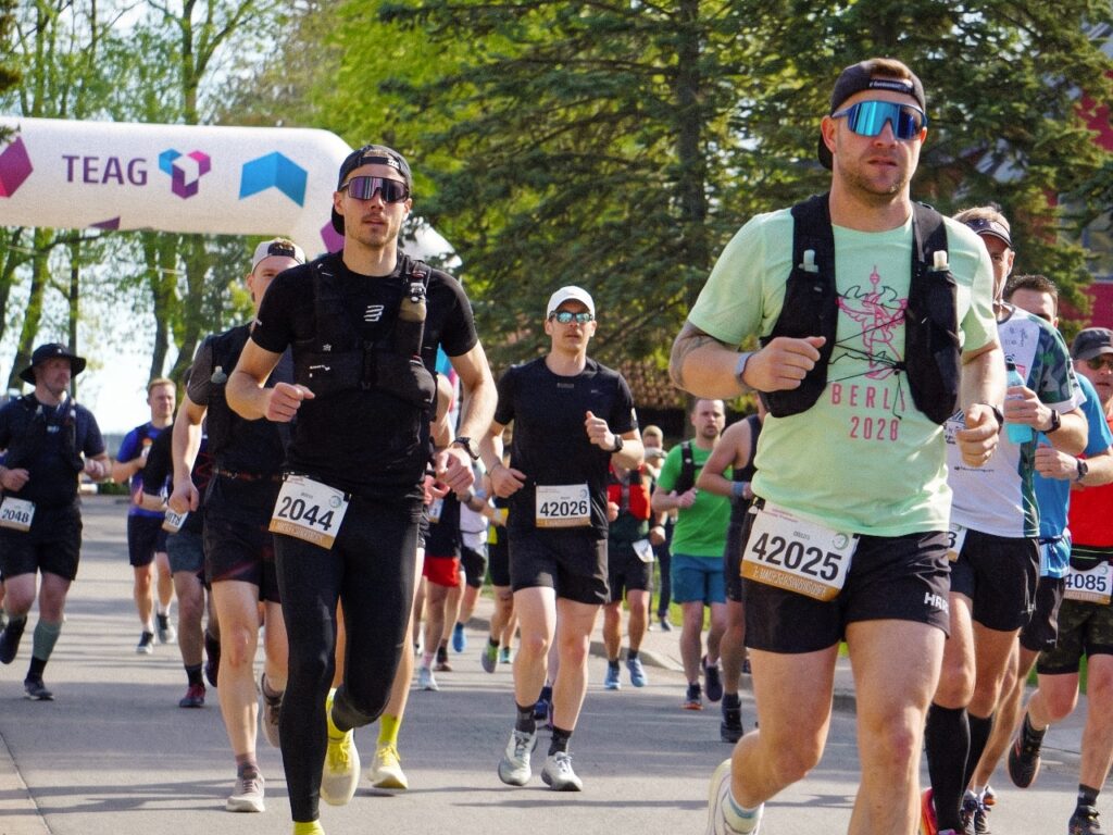 Eine Gruppe Läufer startet unter dem TEAG-Startbogen in Sophienhof. Die Männer tragen Sportkleidung, Sonnenbrillen, Mützen und teils Trinkrucksäcke. Sichtbar sind unter anderem die Startnummern 2044, 42025, 42026, 42048 und 4085. Ein Läufer trägt ein hellgrünes Shirt mit dem Aufdruck „BERLIN 2020". Im Hintergrund stehen Frühlingsbäume entlang der Straße.