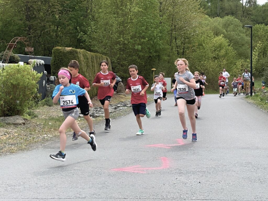 Mehrere Kinder laufen auf einer Asphaltstraße. Vorne ein Mädchen mit pinkem Stirnband in blauem T-Shirt, daneben Jungen in roten Shirts und ein Mädchen in grauem Shirt. Sie tragen Startnummern im 2000er-Bereich (unter anderem 2531, 2574, 2630). Auf der Straße sind pinkfarbene Pfeile als Streckenmarkierung aufgesprüht. Im Hintergrund Sträucher und frisches Frühlingsgrün.