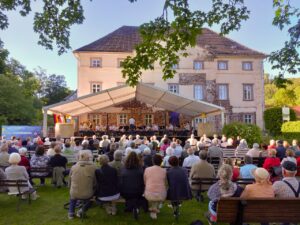 Ein Blasorchester in Uniform spielt unter einem weißen Festzelt vor einem alten Gutshaus. Ein Dirigent steht mit dem Rücken zum Publikum vor den Musikern. Davor sitzen viele Menschen dicht an dicht auf Bänken und schauen zur Bühne. Es ist später Nachmittag oder früher Abend, der Himmel ist blau. Im Vordergrund hängen Zweige eines Ahornbaums ins Bild.