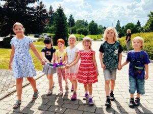 Sieben Kinder gehen bei Sonnenschein &uuml;ber einen gepflasterten Weg. Sie tragen gemeinsam einen Stapel bunter Bilder mit einer lila Schleife. Im Hintergrund sind Wiesen, B&auml;ume und ein Parkplatz zu sehen.