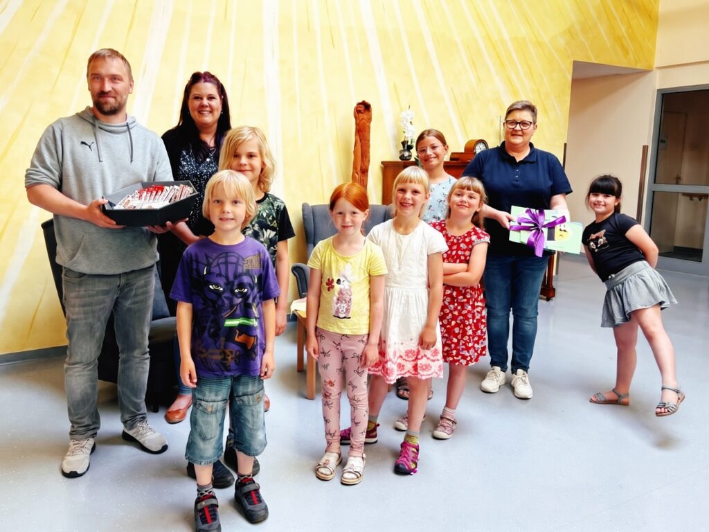 Gruppenfoto in einem hellen Raum mit gelber Wand: Kinder der Evangelischen Grundschule Nordhausen stehen mit Erwachsenen zusammen. Ein Mann hält eine Schale mit Süßigkeiten, eine Frau hält die geschenkten Bilder mit lila Schleife.