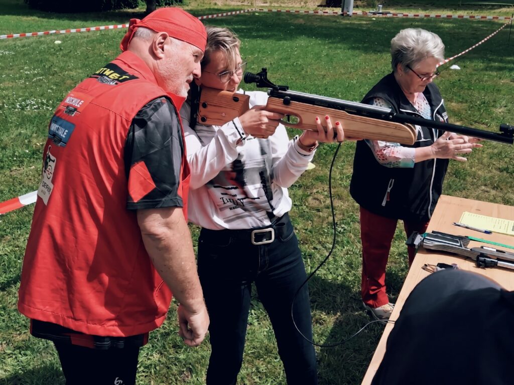 Ein Trainer in rotem Shirt und rotem Kopftuch hilft einer Frau, ein hölzernes Lasergewehr anzulegen und zu zielen. Rechts steht eine Helferin in Weste am Schießstand.