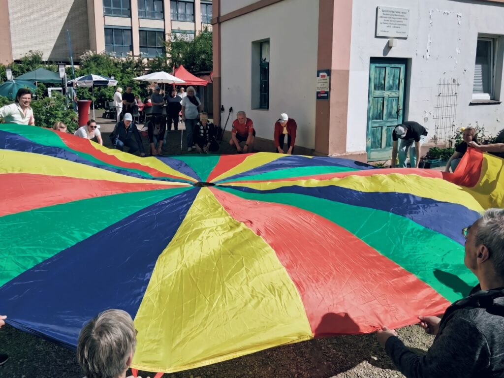 Mehrere Personen halten im Kreis ein großes, buntes Schwungtuch in Regenbogenfarben und bewegen es gemeinsam. Im Hintergrund ein Gebäude und Pavillons des Aktivtags.