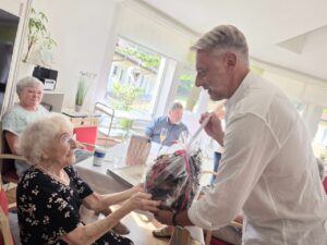 Erika Schirmer sitzt an ihrem 100. Geburtstag an einer festlich gedeckten Tafel im Sonnenhof Ilfeld. Sie trägt ein dunkelblaues Kleid mit Blütenmuster und eine Perlenkette. Bürgermeister Stephan Klante überreicht ihr einen in Folie verpackten Blumenkorb mit roter Schleife. Im Hintergrund sitzen Bewohner und Betreuer am Tisch. Durch große Fenster fällt Sommerlicht in den hellen Raum.