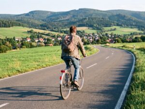 Eine Person faehrt mit einem gewoehnlichen Fahrrad in Alltagskleidung auf einer ruhigen Landstrasse im Harz. Es ist Tag, das Wetter ist freundlich. Das Gesicht ist nicht erkennbar. KI.