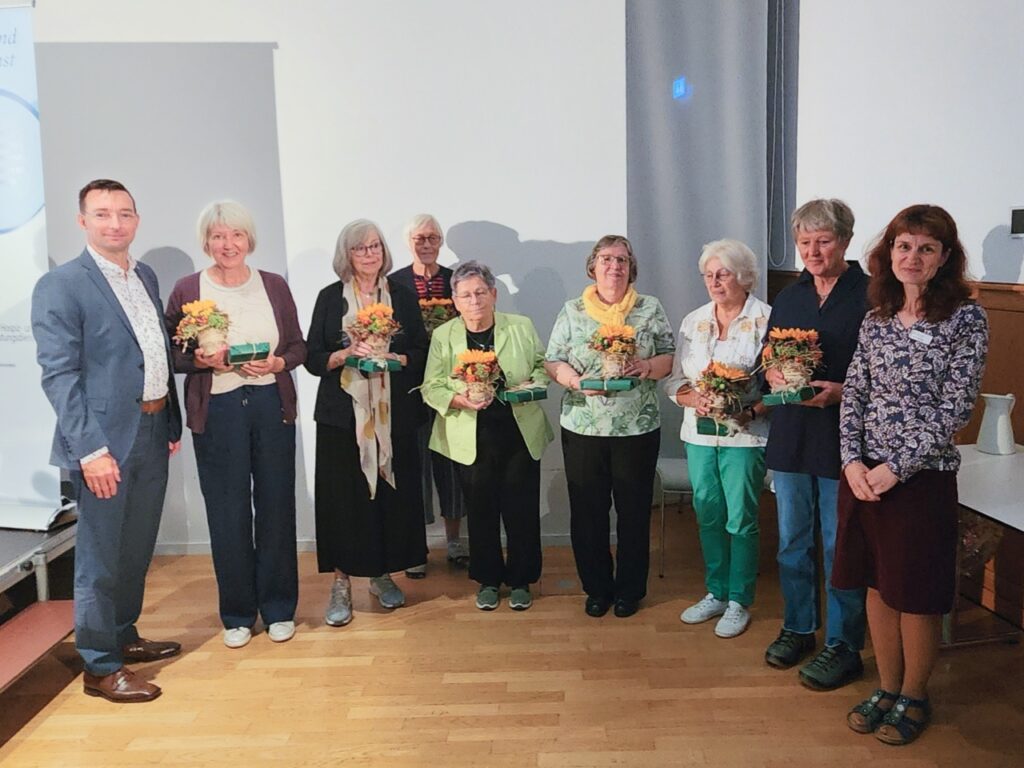 Neun Personen stehen nebeneinander im Ratssaal des Bürgerhauses Nordhausen. Sieben Frauen in der Mitte halten kleine Sonnenblumentöpfe und Präsente in den Händen. Links steht ein Herr im Sakko neben einem Roll-up-Banner des Hospizdienstes, rechts Koordinatorin Judith Roscher.