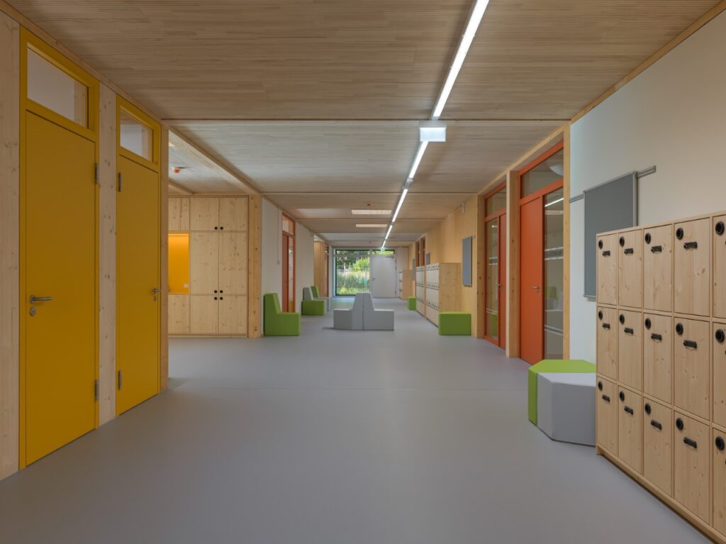 Heller Holzkorridor mit gelben und orangen Türen, Holzgarderoben und grünen Sitzblöcken. Am Ende des Flurs Glas und Bäume.