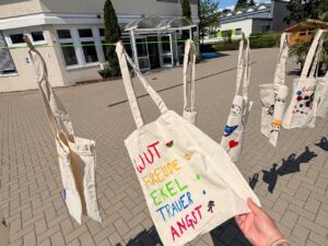 Mehrere selbst bemalte Stoffbeutel h&auml;ngen an einer gr&uuml;nen W&auml;scheleine. Im Vordergrund h&auml;lt eine Hand einen Beutel mit den W&ouml;rtern Wut, Freude, Ekel, Trauer und Angst, jedes in einer anderen Farbe. Dahinter sind weitere Beutel mit Zeichnungen zu sehen, darunter ein Herz und ein Gesicht.