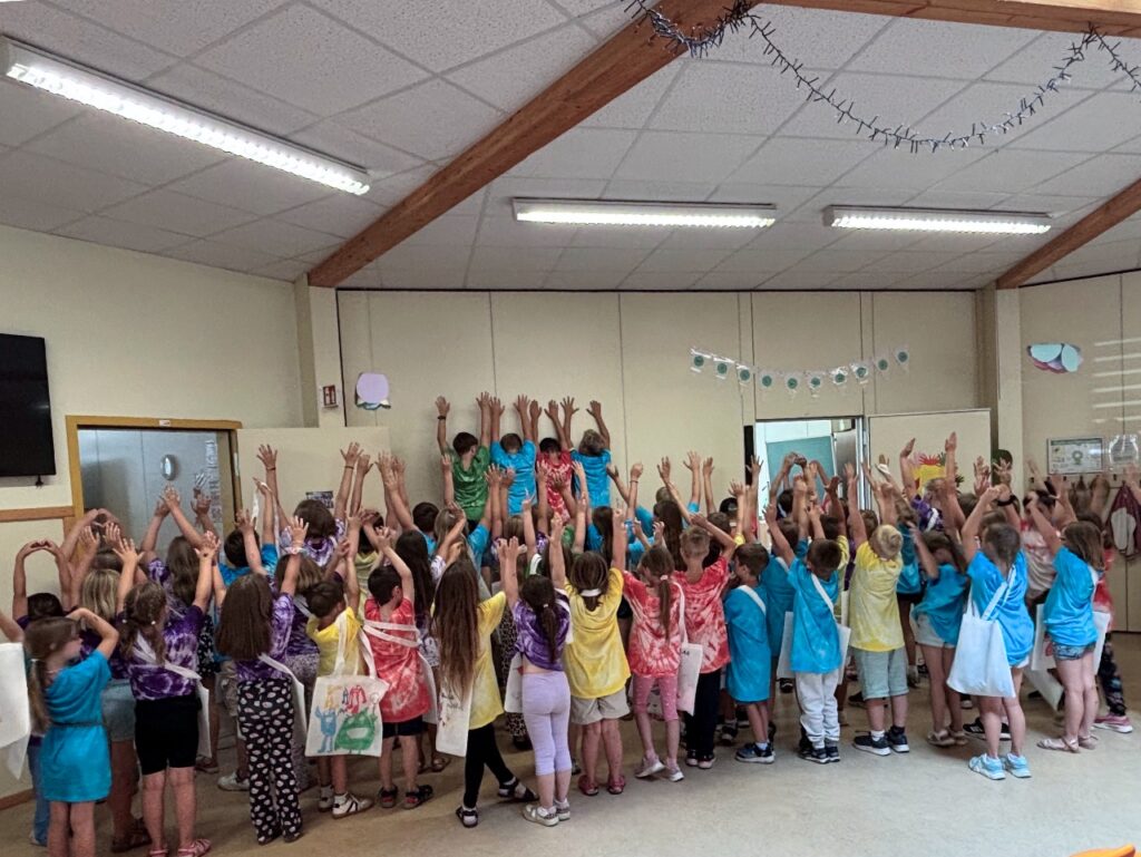 Eine große Gruppe Grundschulkinder steht in einer hellen Halle mit dem Rücken zur Kamera und die Arme nach oben. Die Kinder tragen bunte Batik-Shirts, viele haben selbst bemalte Stoffbeutel über der Schulter. An der Wand hängt eine weiße Wimpelkette.