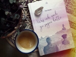 Ein wei&szlig;es Buch mit der Aufschrift &bdquo;Tim Wache: Was ich noch zu sagen h&auml;tte&hellip;&ldquo; und einer Zeichnung zweier Menschen liegt neben einer Tasse Kaffee, einem Bergkristall und einer Topfpflanze auf einem hellen Untergrund.