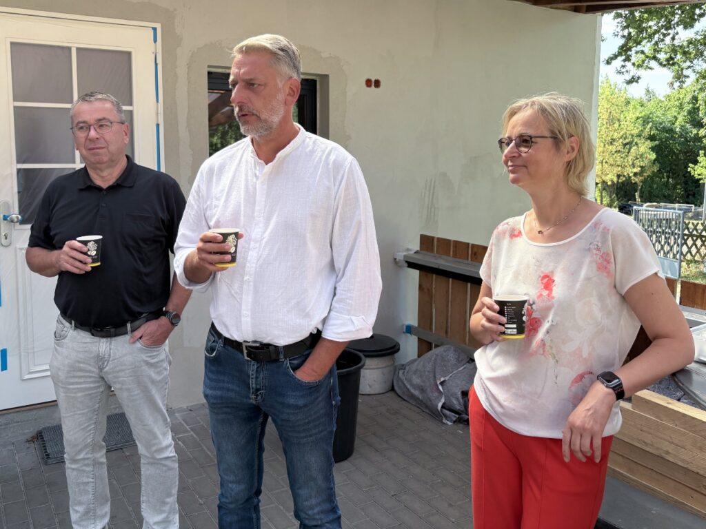 Drei Personen mit Kaffeebechern vor dem frisch verputzten Sportlerheim: Andreas Schmidt vom Bauamt, Bürgermeister Stephan Klante und Finanzministerin Katja Wolf.