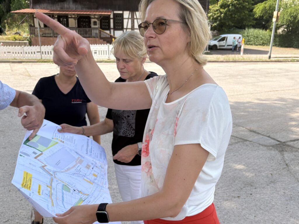 Finanzministerin Katja Wolf hält einen Bauplan in der Hand und zeigt mit dem Arm die Richtung. Zwei Frauen schauen auf den Plan. Im Hintergrund stehen ein Fachwerkhaus und ein Transporter.
