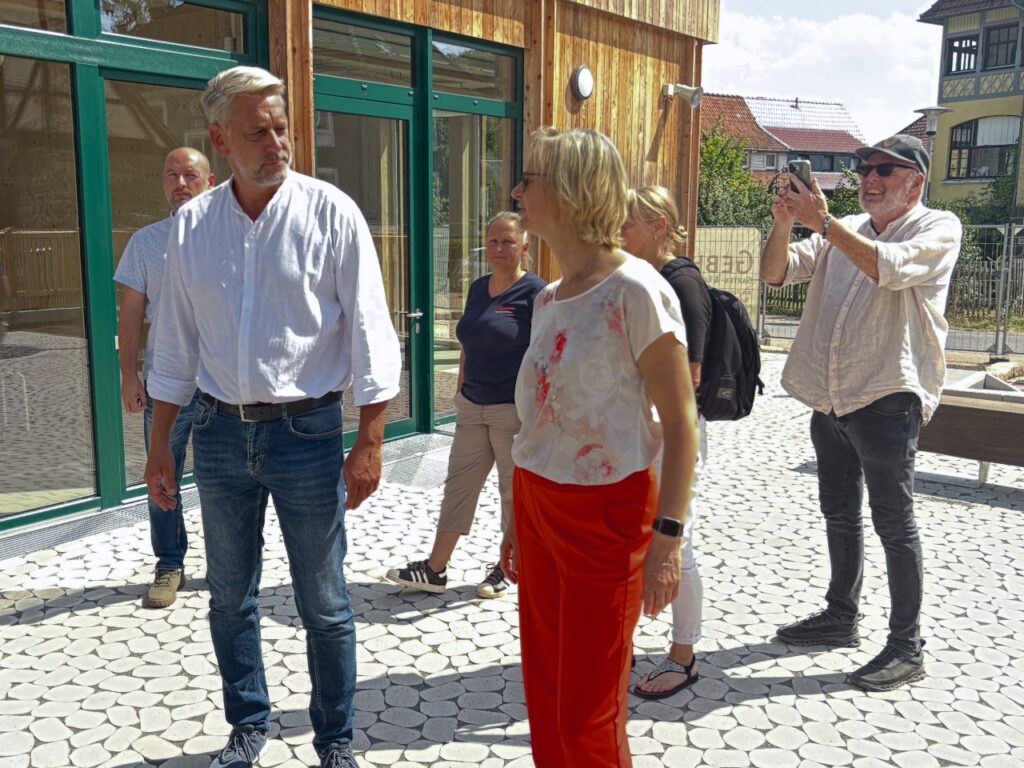 Bürgermeister Stephan Klante und Finanzministerin Katja Wolf vor dem neuen Holzgebäude der Grundschule in Ilfeld. Ein Mann fotografiert die beiden mit dem Handy.