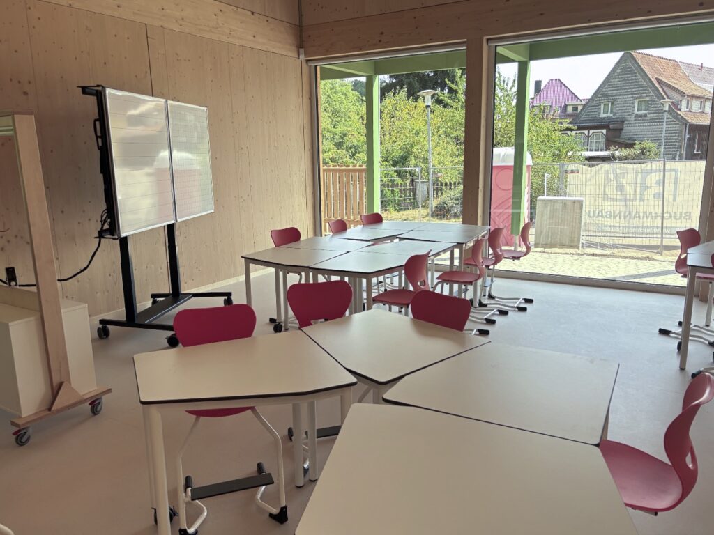 Heller Klassenraum mit Holzwänden, weißen Tischen und roten Stühlen. An der Wand steht eine fahrbare digitale Tafel. Große Fenster geben den Blick nach draußen frei.