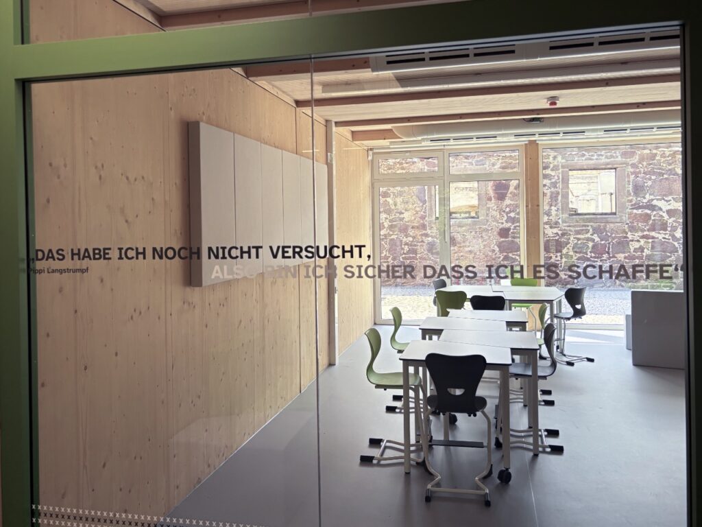 Klassenraum hinter einer Glaswand mit Tischen und grünen Stühlen. Auf dem Glas steht ein Zitat von Pippi Langstrumpf: „Das habe ich noch nicht versucht, also bin ich sicher, dass ich es schaffe."