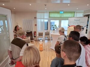Der kleine orangefarbene Empathie-Roboter Paul steht auf dem Holzboden eines hellen Aufenthaltsraums im Sonnenhof. Besucher stehen im Halbkreis um ihn herum. Rechts steht ein Aufsteller mit der Aufschrift „Navel: der Empathie-Roboter in der Pflege".