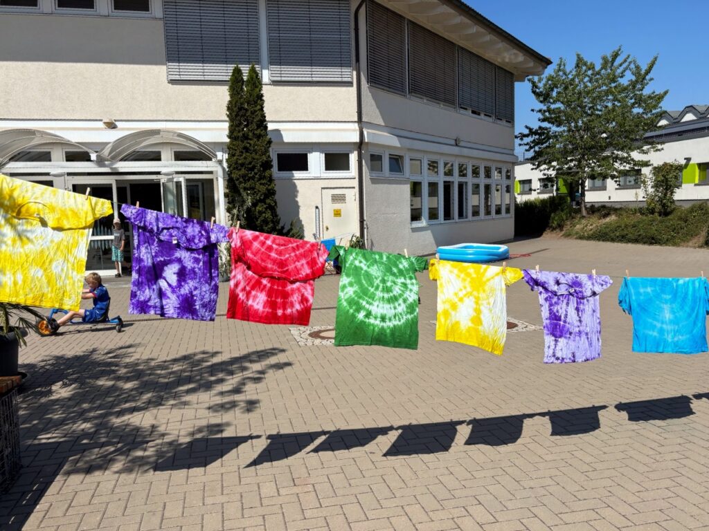 Sieben bunte Batik-T-Shirts hängen auf einer Wäscheleine auf einem gepflasterten Schulhof. Die Shirts sind gelb, lila, rot, grün, helllila und türkis, teils mit Spiralmuster. Im Hintergrund steht ein helles Schulgebäude mit großen Fenstern, rechts ein kleines blaues Planschbecken.