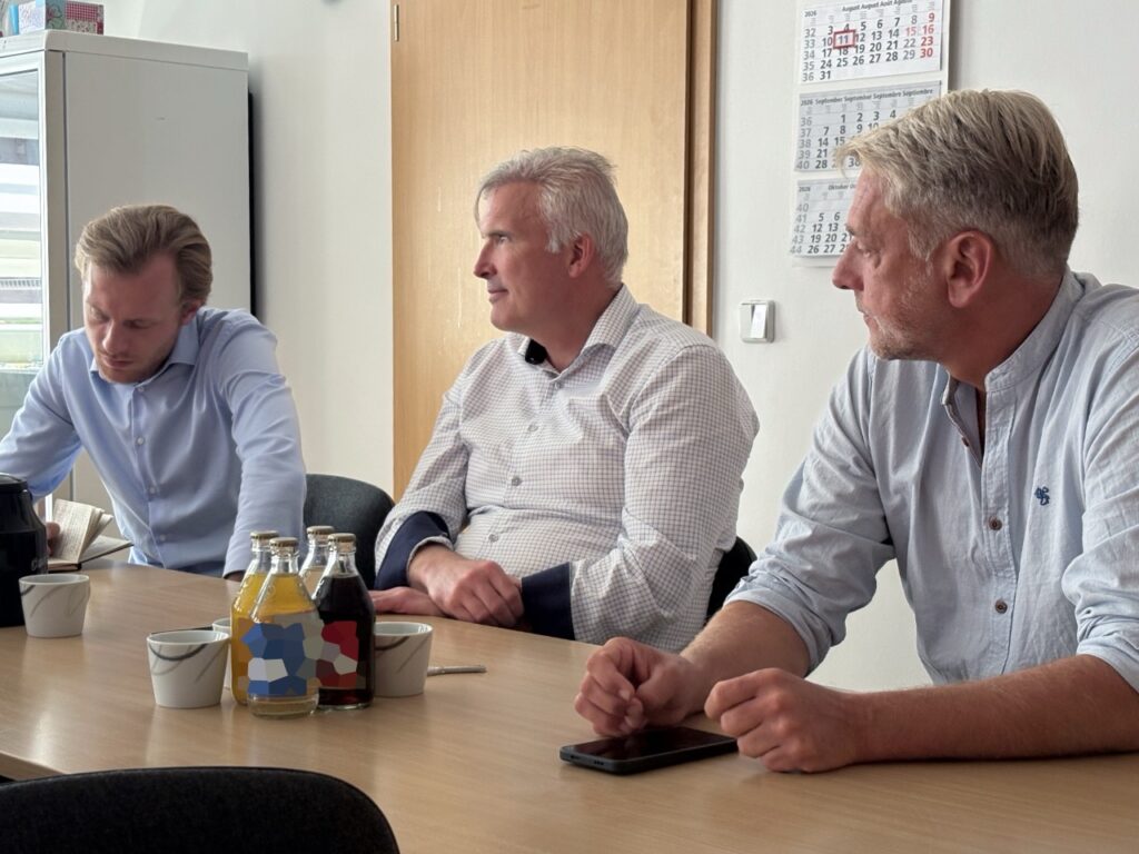 Drei Männer sitzen an einem hellen Holztisch in einem Besprechungsraum. Links notiert ein junger Mann im hellblauen Hemd mit. In der Mitte hört Staatssekretär Andreas Bausewein im weiß karierten Hemd zu, sein linker Arm liegt in einer dunklen Schlinge. Rechts spricht Bürgermeister Stephan Klante im hellblauen Hemd. Auf dem Tisch stehen Saftflaschen, Kaffeetassen und eine Kanne. An der Wand hängt ein Kalender, der 11. August ist rot markiert.