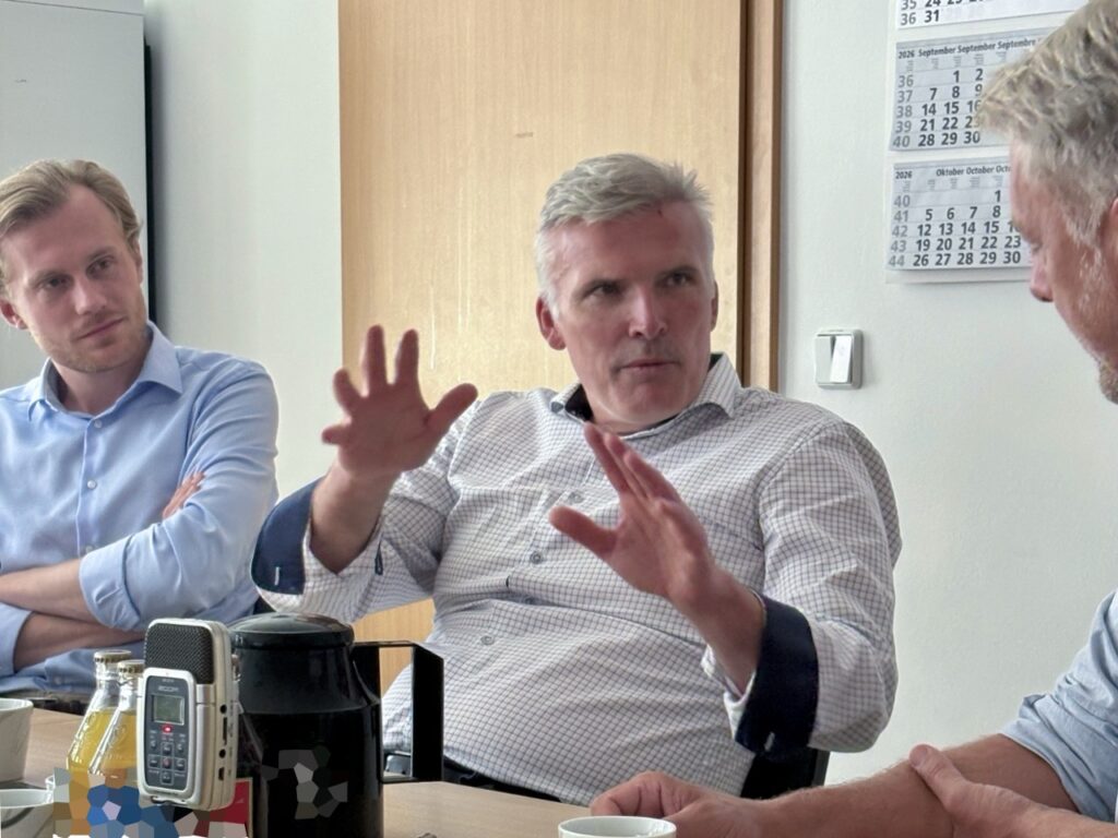 Staatssekretär Andreas Bausewein sitzt am Besprechungstisch und erklärt etwas mit beiden Händen. Er trägt ein weiß kariertes Hemd, sein linker Arm liegt in einer dunklen Schlinge. Links hört ein junger Mann im hellblauen Hemd mit verschränkten Armen zu, rechts im Anschnitt ein weiterer Gesprächsteilnehmer. Auf dem Tisch stehen ein Aufnahmegerät, eine Kaffeekanne und Getränke.