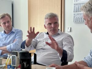 Staatssekret&auml;r Andreas Bausewein sitzt am Besprechungstisch und erkl&auml;rt etwas mit beiden H&auml;nden. Er tr&auml;gt ein wei&szlig; kariertes Hemd, sein linker Arm liegt in einer dunklen Schlinge. Links h&ouml;rt ein junger Mann im hellblauen Hemd mit verschr&auml;nkten Armen zu, rechts im Anschnitt ein weiterer Gespr&auml;chsteilnehmer. Auf dem Tisch stehen ein Aufnahmeger&auml;t, eine Kaffeekanne und Getr&auml;nke.