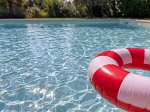 Nahaufnahme von klarem Wasser in einem Freibad. Die Sonne glitzert auf kleinen Wellen. Am Bildrand schwimmt ein rot-weißer Schwimmring, im Hintergrund sind unscharfe Bäume zu sehen. KI.