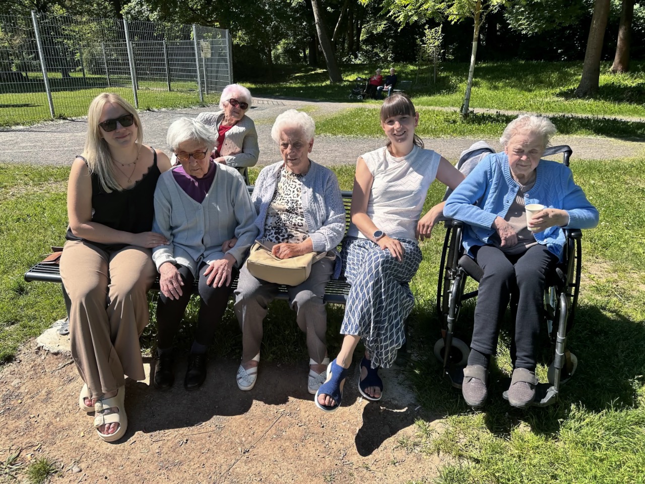 Zwei Mitarbeiterinnen sitzen mit vier älteren Frauen auf einer Bank im Park, eine weitere Frau sitzt im Rollstuhl daneben. Alle blicken lächelnd in die Kamera. Es ist ein sonniger Tag, im Hintergrund sind Bäume, ein Weg und eine Wiese zu sehen.