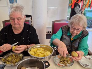 Zwei ältere Frauen sitzen an einem Tisch und schälen gemeinsam Kartoffeln. Vor ihnen stehen Schüsseln mit geschälten Kartoffeln und mit Kartoffelschalen. Die Szene spielt drinnen in einem hellen Raum.