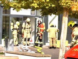Mehrere Feuerwehrleute in beiger Schutzkleidung mit gelben Helmen und Atemschutzger&auml;ten stehen vor dem Glaseingang der Bundesfachschule. &Uuml;ber der T&uuml;r steht &bdquo;K&auml;lte-Klima-Technik&ldquo; mit einer Schneeflocke als Logo. Ein Feuerwehrmann in der Mitte tr&auml;gt eine Atemschutzmaske. Auf dem Boden liegen Schl&auml;uche. Vorn h&auml;ngen herbstliche Bl&auml;tter ins Bild.