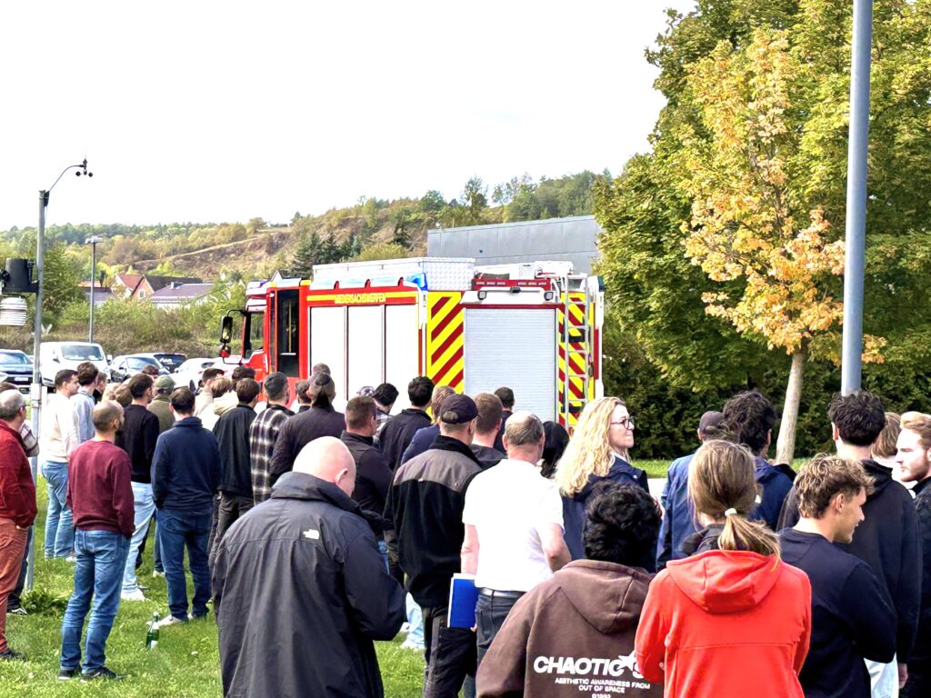 Viele Menschen stehen auf einer Wiese, von hinten zu sehen. Vor ihnen steht ein rotes Löschfahrzeug mit der Aufschrift „Niedersachswerfen“ und Blaulicht. Im Hintergrund liegen ein bewaldeter Hang und ein Baum mit gelb-orangem Laub.