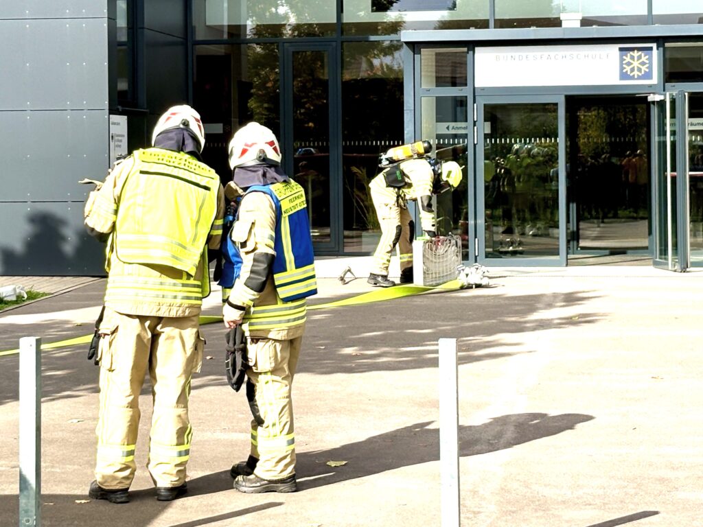 Zwei Feuerwehrleute mit weißen Helmen stehen mit dem Rücken zur Kamera. Einer trägt eine gelbe Weste mit der Aufschrift „Einsatzleiter Harztor“, der andere eine blaue Gruppenführer-Weste. Vor dem Eingang der Schule bereitet ein Feuerwehrmann mit Atemschutzgerät Material vor. Ein gelber Schlauch liegt auf dem Vorplatz.