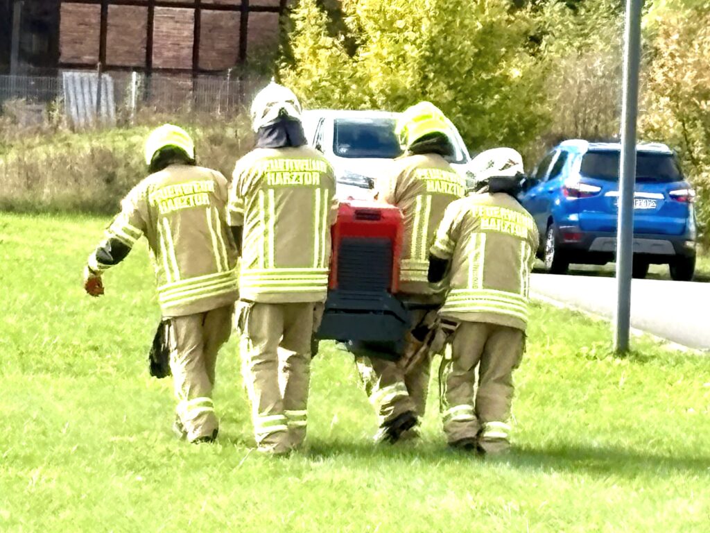 Vier Feuerwehrleute mit Helmen und Jacken mit der Aufschrift „Feuerwehr Harztor“ gehen von hinten gesehen über eine Wiese. Gemeinsam tragen sie eine große rote Pumpe. Rechts verläuft eine Straße mit zwei Autos.