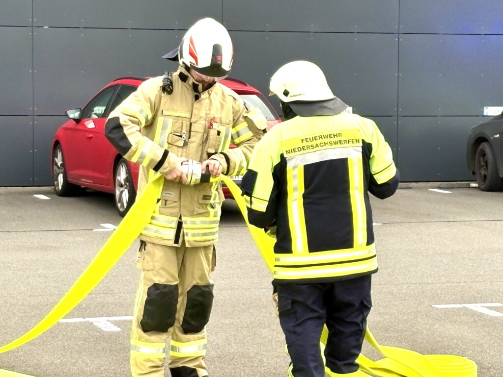 Zwei Feuerwehrleute stehen auf einem Parkplatz vor einer dunklen Wand. Einer verbindet zwei gelbe Schläuche mit einer Kupplung. Der andere trägt eine Jacke mit der Aufschrift „Feuerwehr Niedersachswerfen“. Dahinter parkt ein rotes Auto.