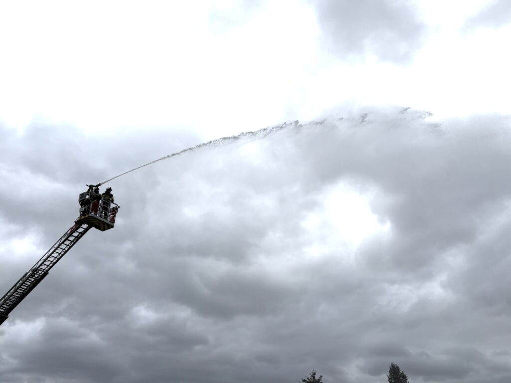 Eine Drehleiter ragt schräg in einen grauen, wolkigen Himmel. Im Korb stehen zwei Feuerwehrleute. Aus dem Rohr schießt ein langer Wasserstrahl nach rechts.
