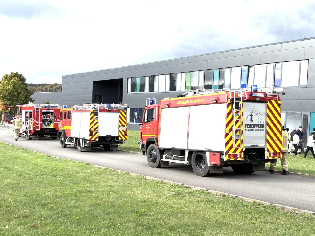 Drei rote Feuerwehrfahrzeuge stehen hintereinander auf einem Weg neben dem dunklen Gebäude der Bundesfachschule. Auf dem vorderen steht „Neustadt-Osterode“. Links geht ein Feuerwehrmann, der Himmel ist dunkel bewölkt.