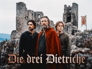 Drei M&auml;nner in schlichter mittelalterlicher Kleidung vor einer steinernen Burgruine bei bedecktem Himmel. In der Mitte ein &auml;lterer Mann mit grauem Bart und rotem Mantel, links und rechts zwei j&uuml;ngere M&auml;nner. Rechts im Hintergrund ein kleines Banner mit Geweih &uuml;ber rot-wei&szlig;em Schachbrett.
