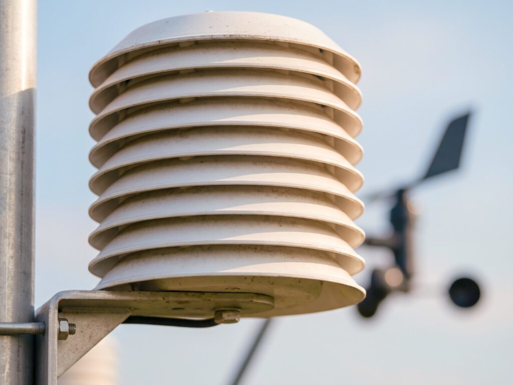 Nahaufnahme einer automatischen Wetterstation: weißer Strahlungsschutz mit Lamellen, Mast und Windmesser unscharf im Hintergrund. KI.
