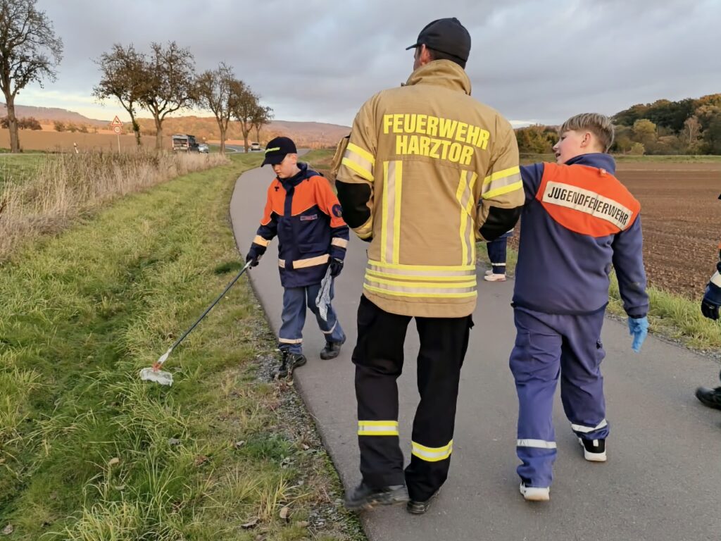 Von hinten: ein Erwachsener in Jacke mit Aufschrift Feuerwehr Harztor und Kinder der Jugendfeuerwehr auf einem Weg neben einem Acker. Ein Kind holt mit dem Greifer etwas aus dem Gras.