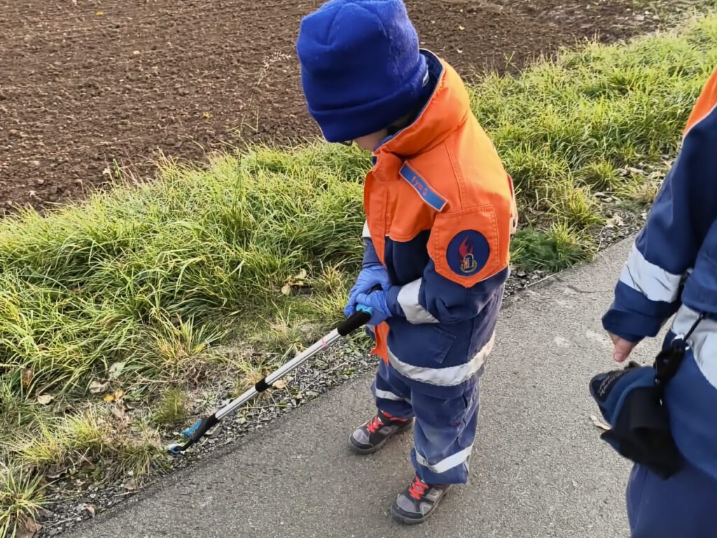 Ein Kind der Jugendfeuerwehr in blau-oranger Uniform und blauer Mütze beugt sich mit Greifer und Handschuhen über den Wegrand. Neben dem Weg ein Acker.
