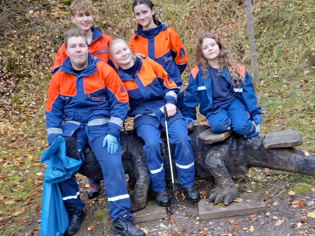 Fünf Kinder der Jugendfeuerwehr posieren lächelnd an einem großen Holzspielgerät im Herbstlaub. Ein Kind hält einen blauen Müllsack.