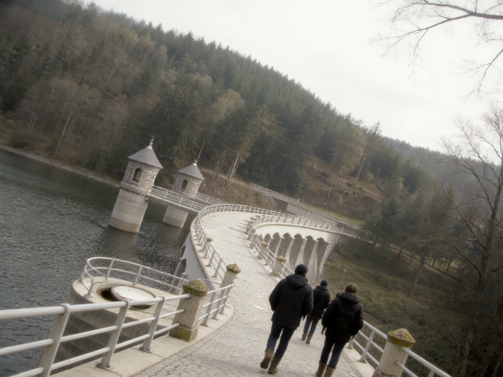 Drei Wandernde auf der Staumauer der Talsperre Neustadt, links das Staubecken mit Entnahmetürmen, rechts bewaldeter Hang. KI-Symbolbild