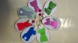 Bastelarbeit in Blumenform: Acht ovale Anhänger aus Pappe sind kreisförmig angeordnet und jeweils mit bunten Osterhasen aus Glitzerpapier verziert. In der Mitte verbinden kleine künstliche Blüten die Anhänger. Die Hasen sind in verschiedenen Farben gestaltet – darunter Grün, Pink, Silber, Blau und Rot. Die Dekoration liegt auf einem hellen Untergrund.
