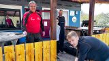 **ALT-Text:** Vier Männer und ein Kind arbeiten bei Sonnenschein gemeinsam an der Instandsetzung eines Holzgeländers vor dem Vereinsheim des SV Ilfeld. Im Hintergrund ist das Vereinslogo zu erkennen, die Stimmung wirkt freundlich und engagiert.