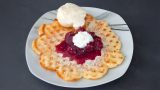 Eine goldbraun gebackene, herzförmige Waffel liegt auf einem weißen Teller; darauf in der Mitte ein Klecks leuchtend roter Kirschsoße mit geschlagener Sahne, rundherum Puderzucker. Oben auf der Waffel ruht eine Kugel Vanilleeis.