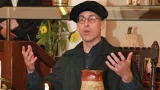 Darsteller in schwarzer Gelehrtenrobe und Barett gestikuliert beim Vortrag; Kerze und Tonkrug im Vordergrund, Blumenarrangement dahinter.
